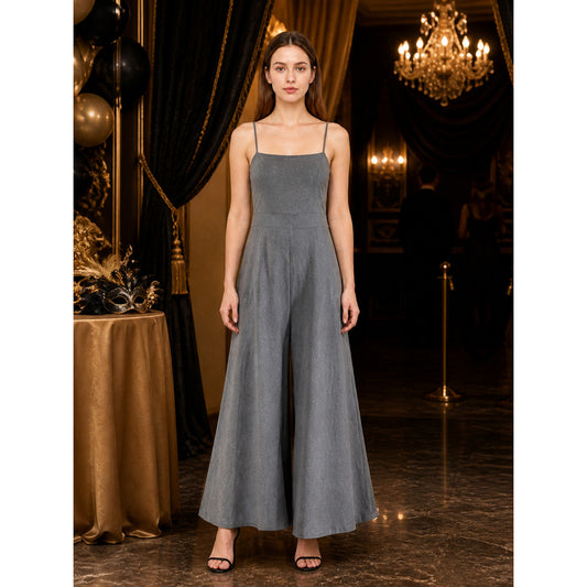 Gray wide-leg jumpsuit with thin shoulder straps and a front center pleat, shown full-length on a model in an indoor evening setting