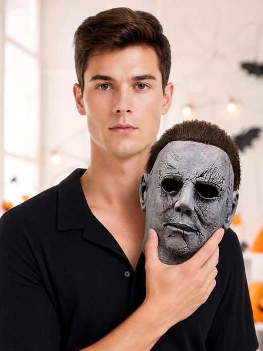 Gray full-face mask with molded scar lines, dark eye openings, and a brown hair-like top held beside a model’s face