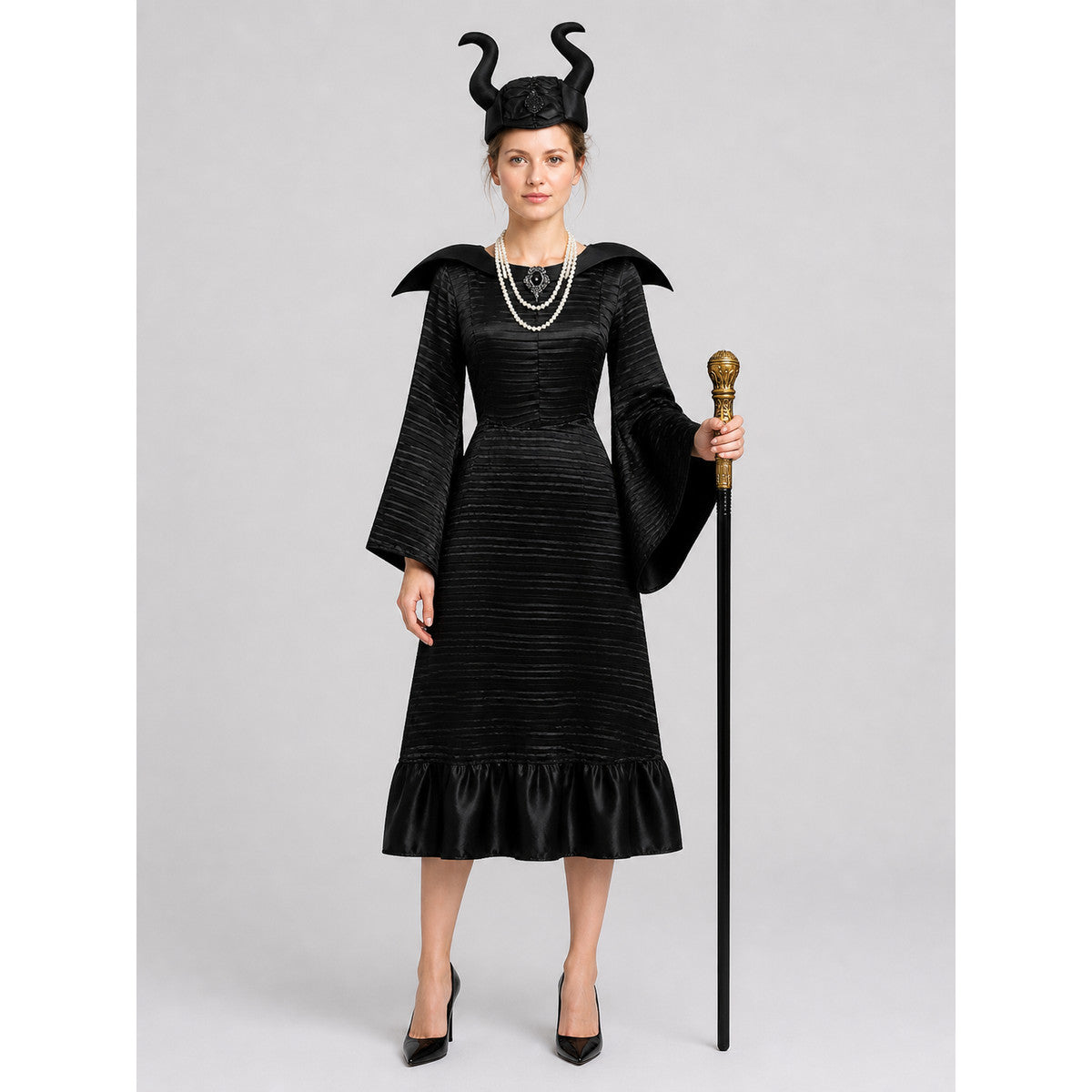 Model wearing a black pleated witch costume: long bell-sleeve dress with pointed collar, horned hat, pearl necklace and gold-knob staff, front view