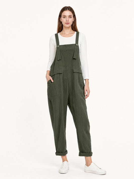 Full-body model wearing dark green textured bib overalls with a chest pocket, side pockets, metal buttons, rolled cuffs, and white sneakers