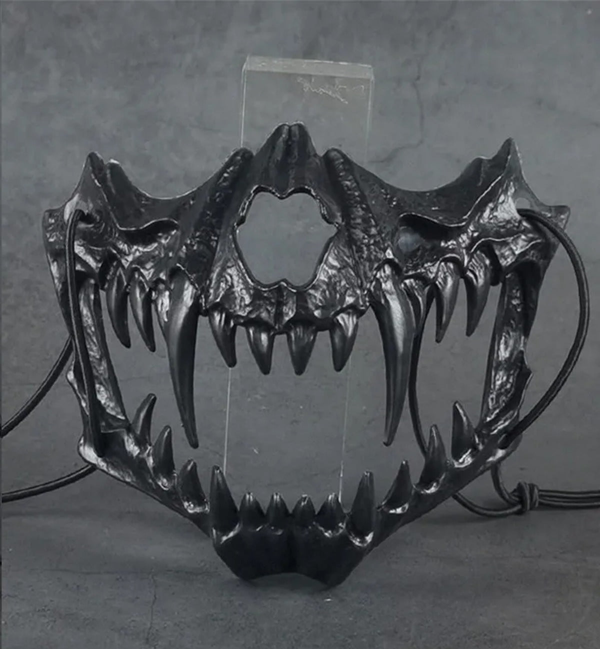 Actual product photo of the black skull jaw half mask with fangs and black cord ties, shown on a clear display stand (stand not included)