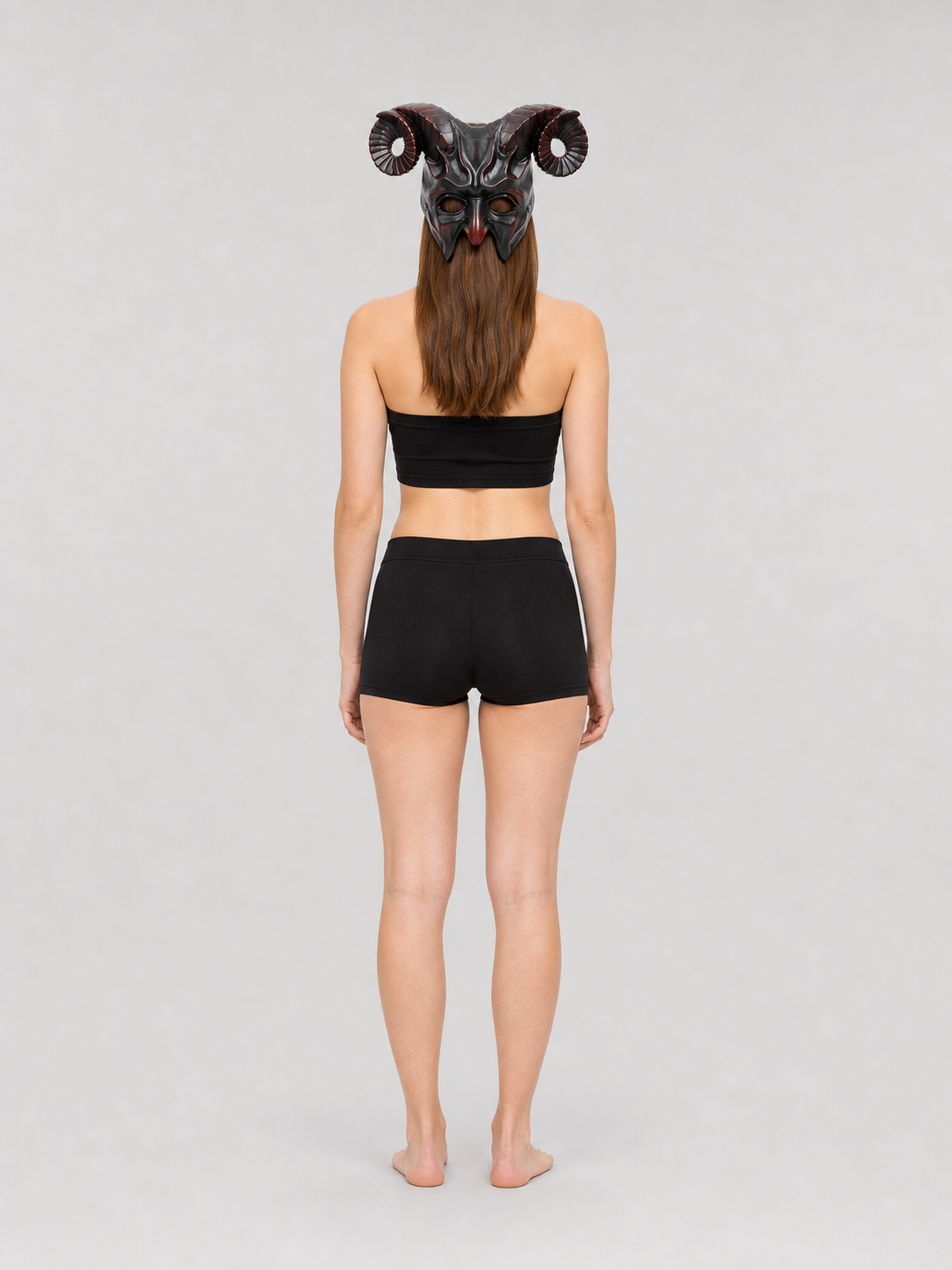 Full-body rear view of a woman wearing the horned mask with curled ridged horns, a black top, and black shorts