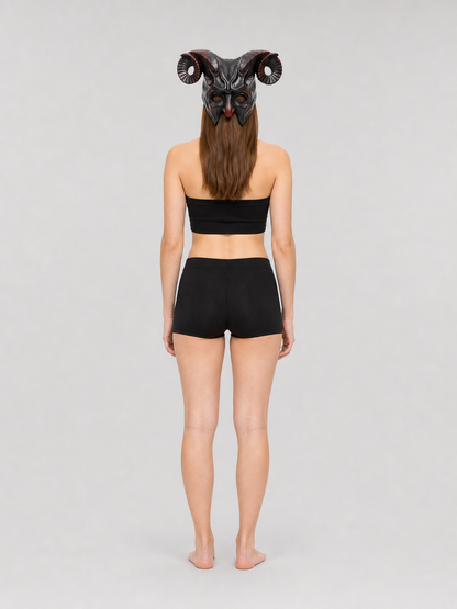Full-body rear view of a woman wearing the horned mask with curled ridged horns, a black top, and black shorts