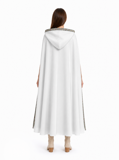 White hooded cape shown from the back on a model, with a plain flowing body and patterned trim around the hood and edges