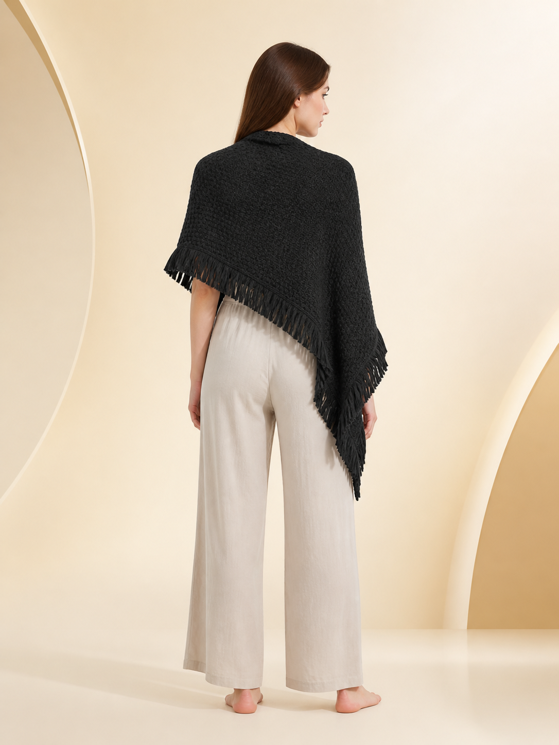 Back view of a black knitted poncho cape on a model, showing the triangular drape and fringe along the lower edges