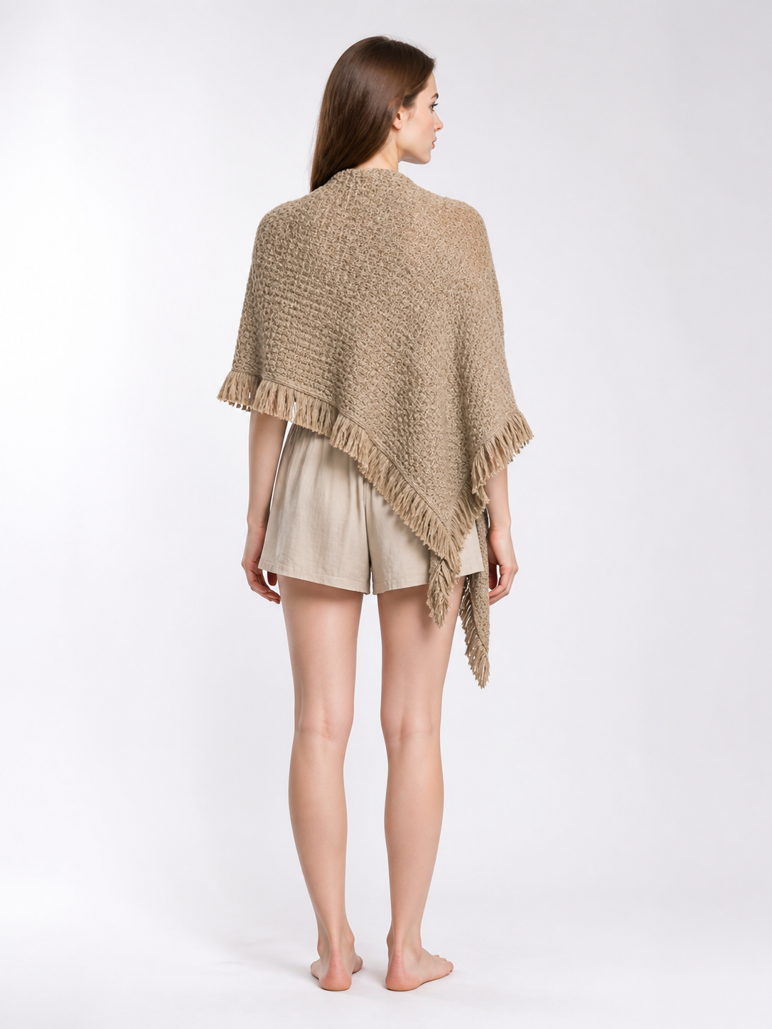 Back view of a beige fringed cape on a model outdoors, showing the textured weave and pointed lower edge