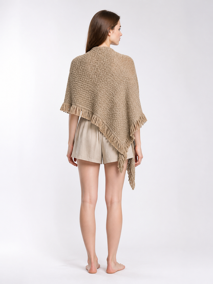 Back view of a beige fringed cape on a model outdoors, showing the textured weave and pointed lower edge