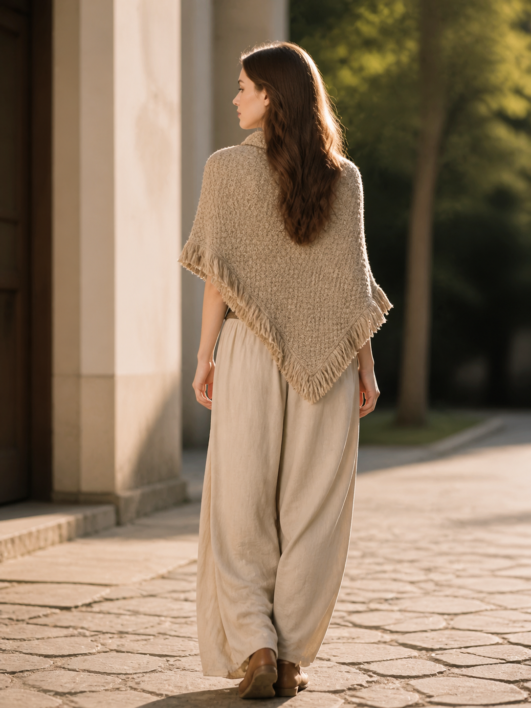 Back view of a beige woven cape with fringed trim, styled with loose light trousers in an outdoor setting