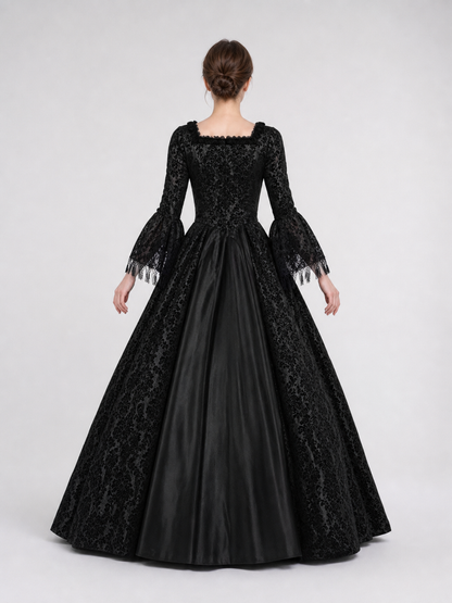 The black dress is designed in the aesthetic of a Victorian-era Gothic costume. The model features a floor-length silhouette, a fitted bodice with a square neckline, and decorative lacing or trim on the front. The long flared sleeves end in