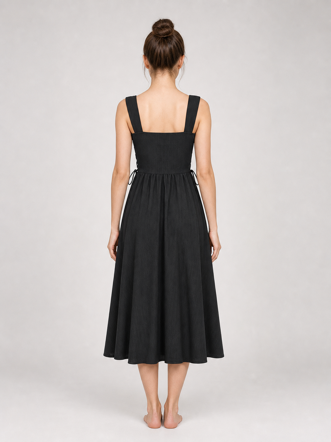 Back view of a black midi dress on a model with hair in a bun, showing wide straps, a square neckline, side ties, and a gathered skirt