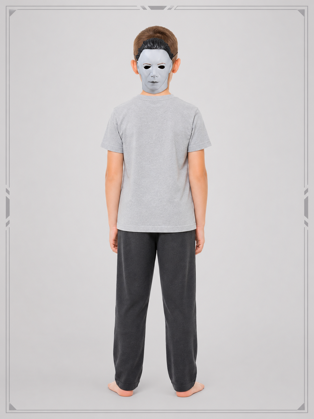 Full-body rear view of a child wearing the white face mask, gray shirt, and dark loose trousers against a gray studio background