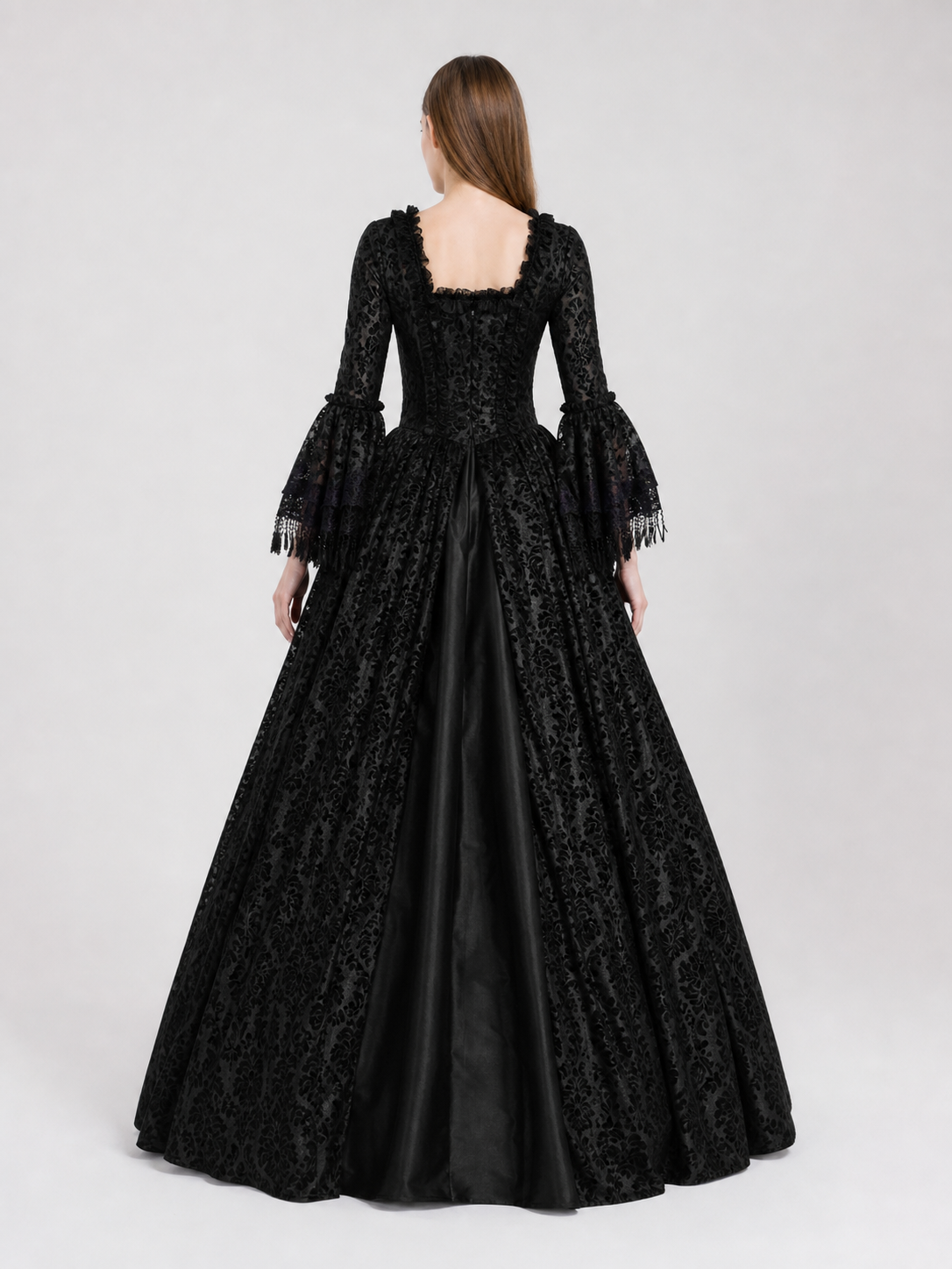 The black dress is designed in the aesthetic of a Victorian-era Gothic costume. The model features a floor-length silhouette, a fitted bodice with a square neckline, and decorative lacing or trim on the front. The long flared sleeves end in