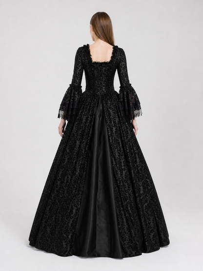 The black dress is designed in the aesthetic of a Victorian-era Gothic costume. The model features a floor-length silhouette, a fitted bodice with a square neckline, and decorative lacing or trim on the front. The long flared sleeves end in