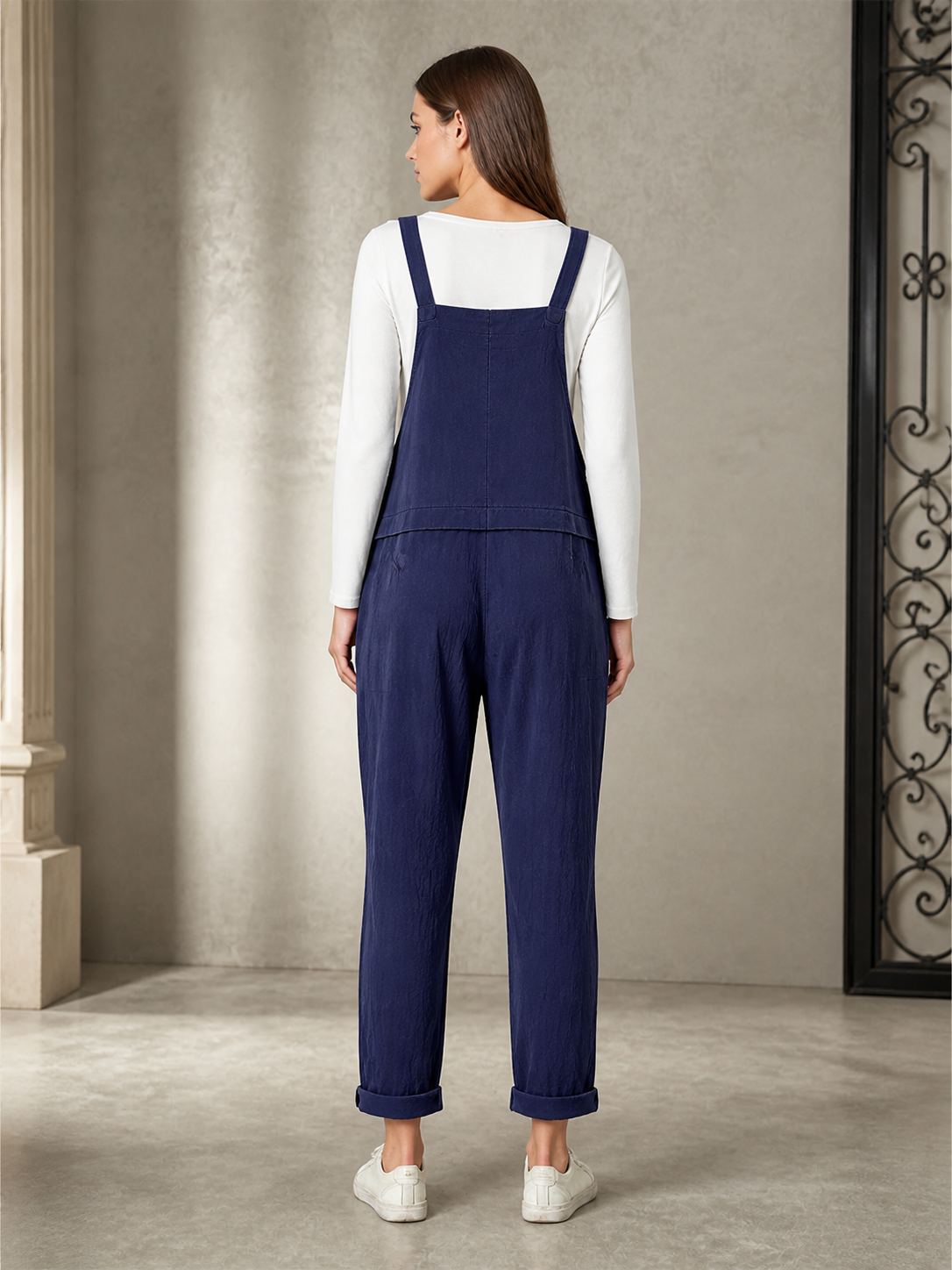Back view of navy blue bib overalls over a white shirt, showing the back bib, rear pockets, loose legs, and cuffed hems
