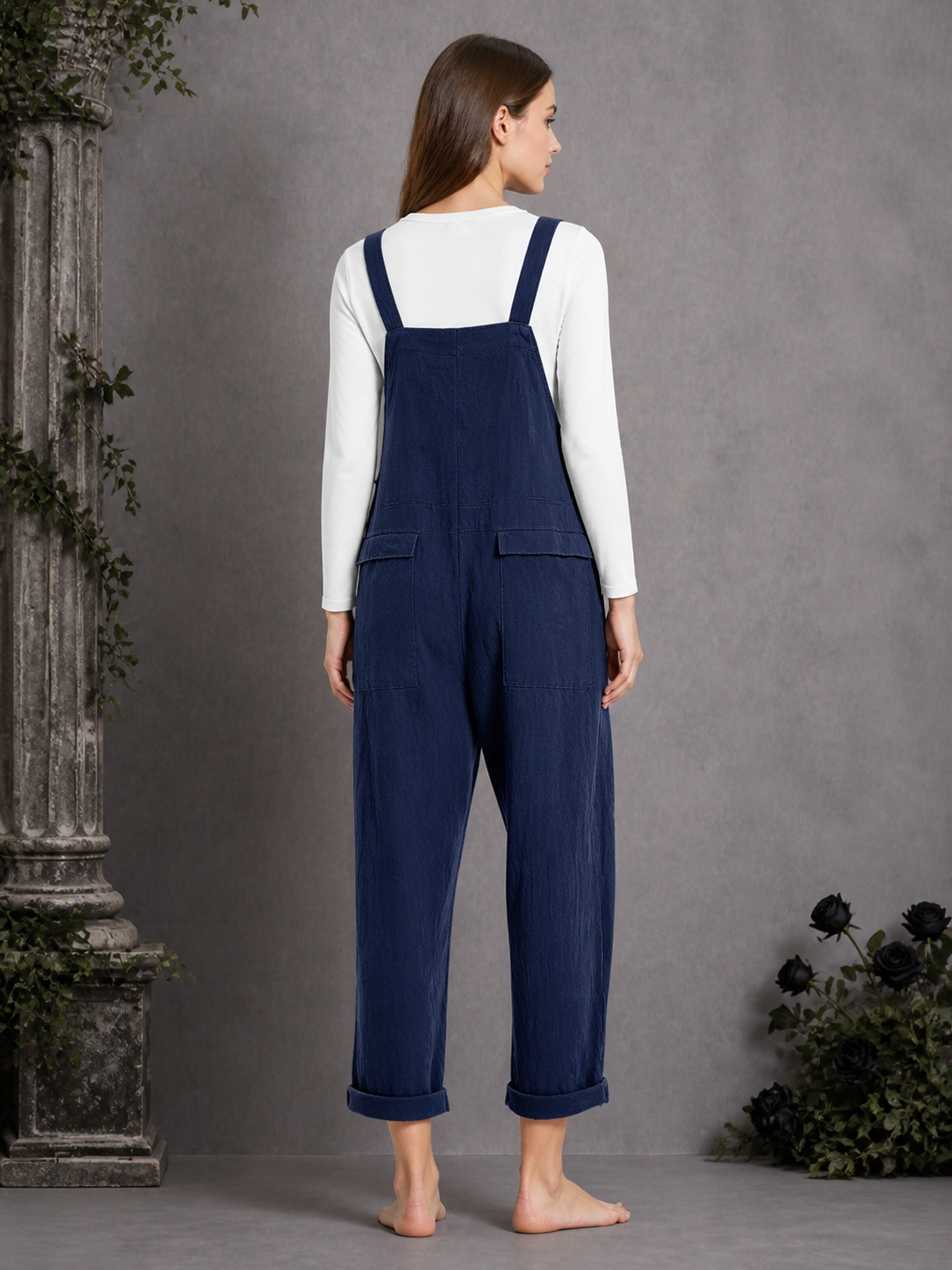 Back view of navy blue bib overalls on a model standing barefoot, showing the back bib panel, rear pockets, and rolled leg hems
