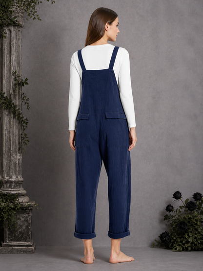 Back view of navy blue bib overalls on a model standing barefoot, showing the back bib panel, rear pockets, and rolled leg hems