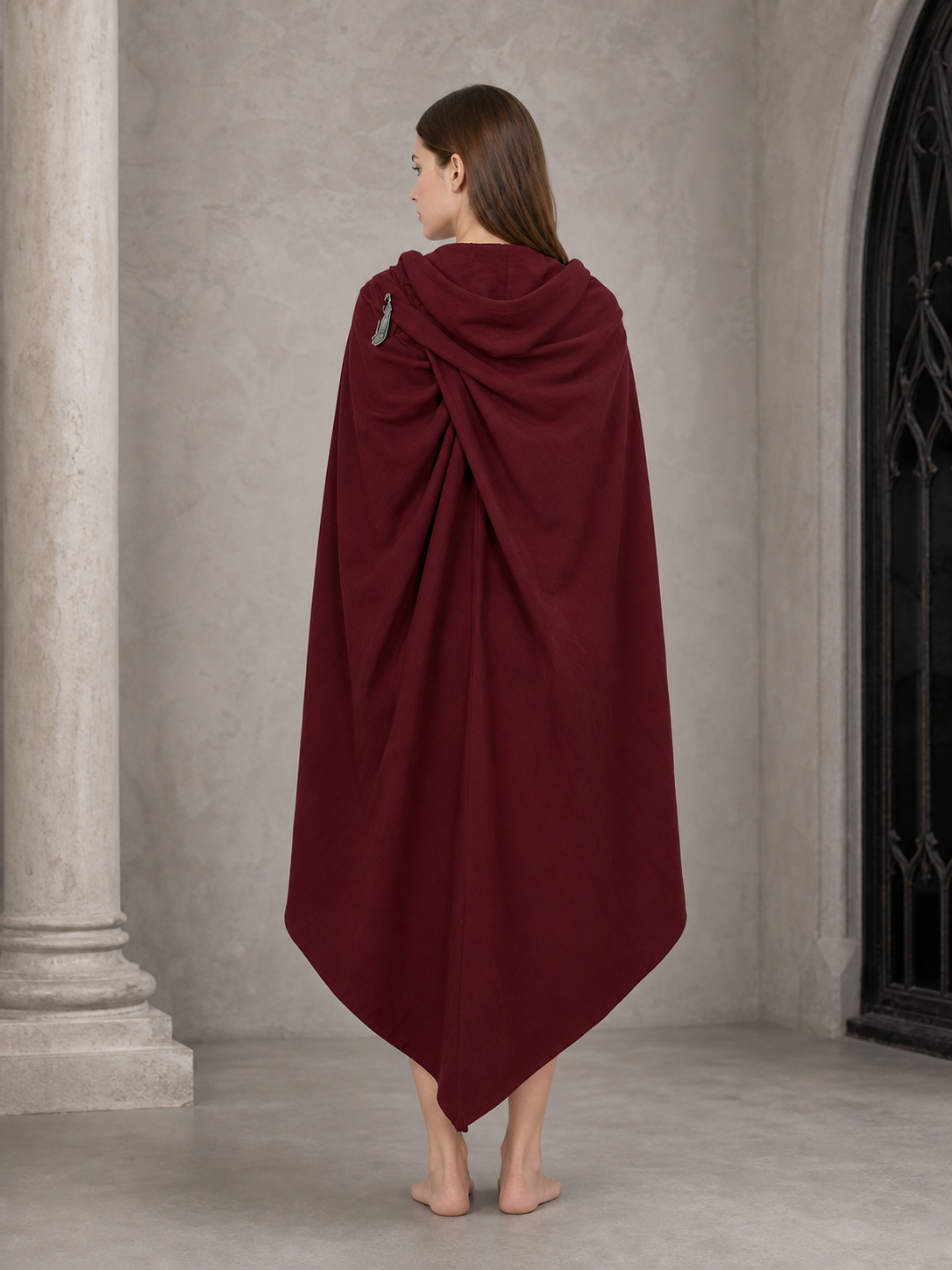 Back view of a burgundy hooded cloak with draped hood, shoulder fastener, and long pointed hem near a stone doorway