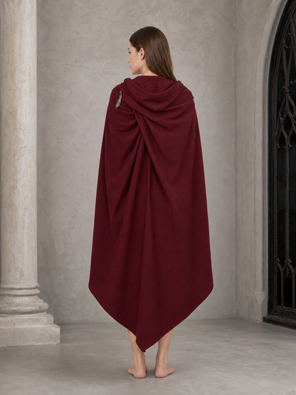 Back view of a burgundy hooded cloak with draped hood, shoulder fastener, and long pointed hem near a stone doorway