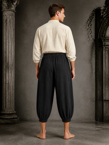 Back view of a man wearing black cropped harem pants with a gathered waistband, loose legs, and ankle cuffs in a stone interior