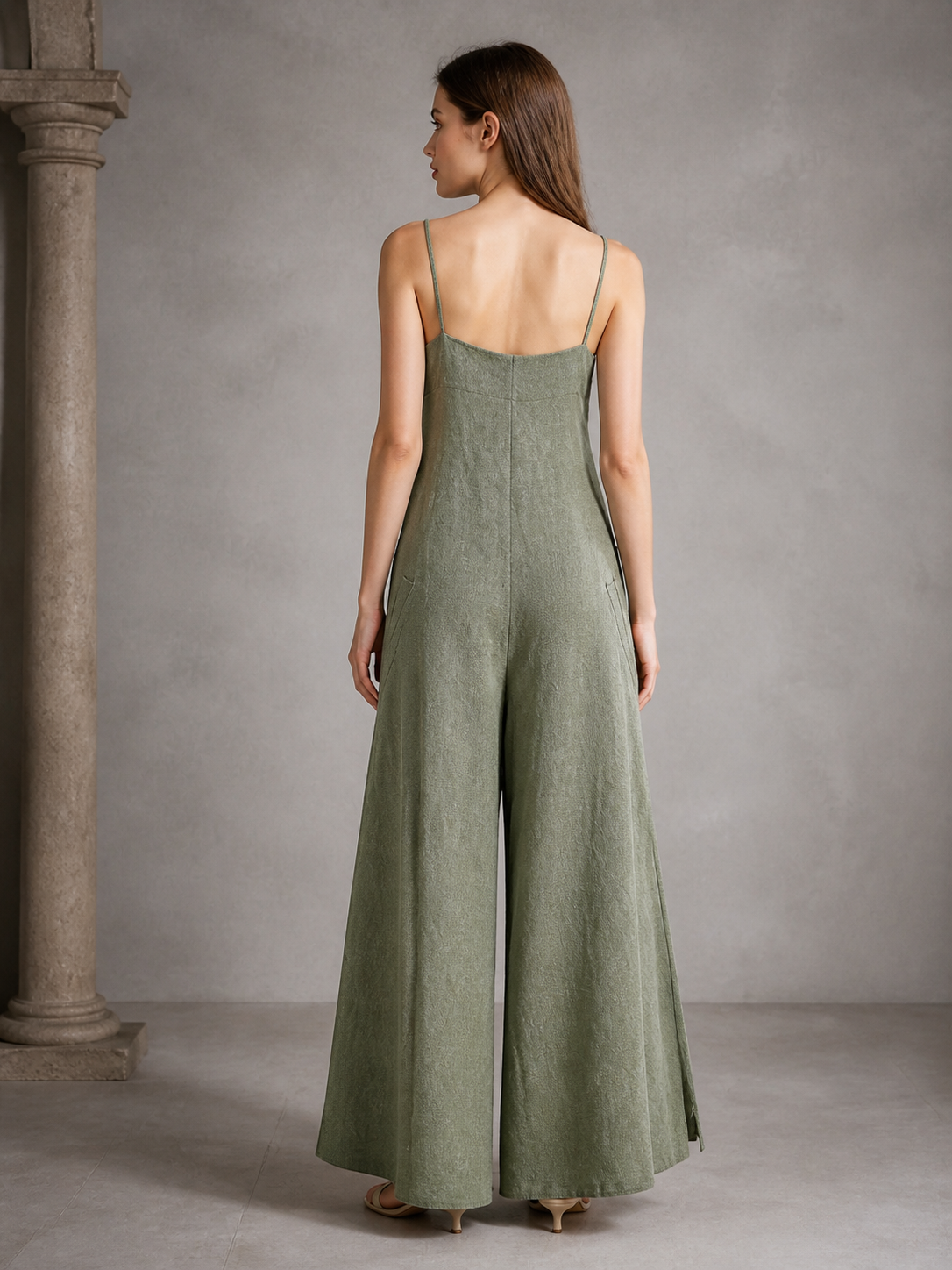 Back view of an olive green wide-leg jumpsuit with thin straps, shown on a model in a gray interior with a column