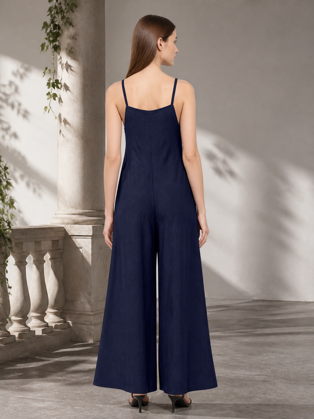 Back view of a navy blue wide-leg jumpsuit with thin straps shown on a model outdoors beside a pale wall