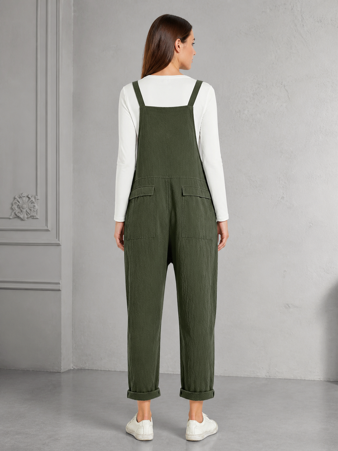 Back view of dark green bib overalls showing the wide back panel, shoulder straps, two rear pockets, rolled cuffs, and white sneakers