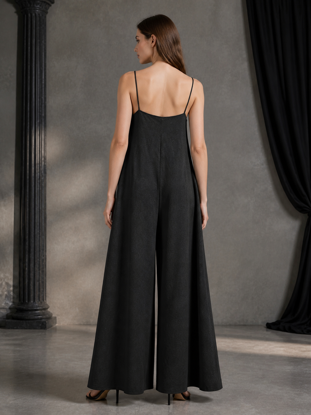 Back view of a black wide-leg jumpsuit with thin straps, shown on a model against a gray wall with black drapery