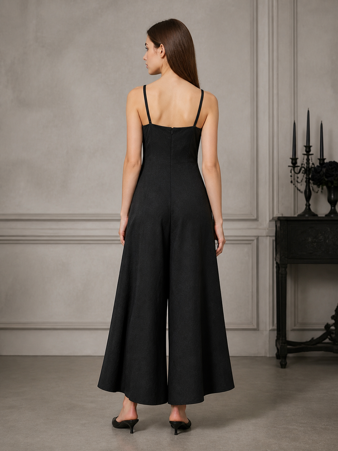 Back view of a black wide-leg jumpsuit with thin straps and a center seam, modeled in a gray paneled room