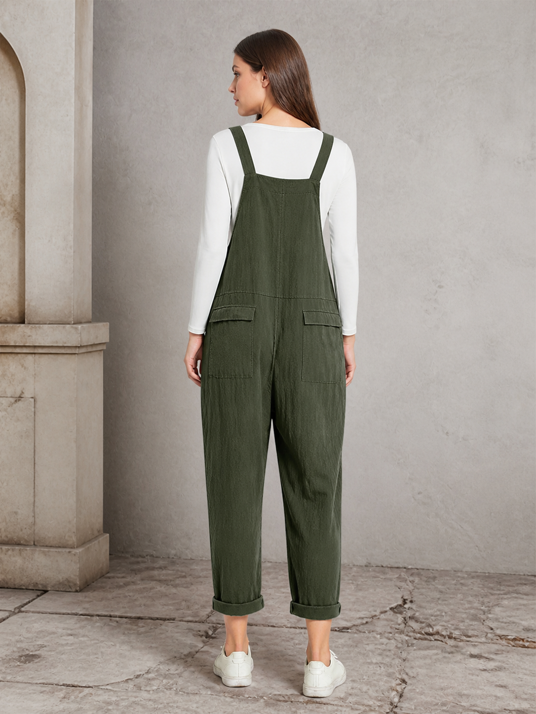 Back view of dark green bib overalls showing the shoulder straps, wide back panel, two rear pockets, and rolled cuffs against a stone interior