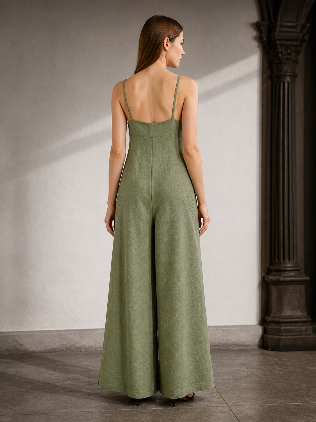 Back view of an olive green wide-leg jumpsuit with thin straps and wide legs, shown on a model in an indoor archway
