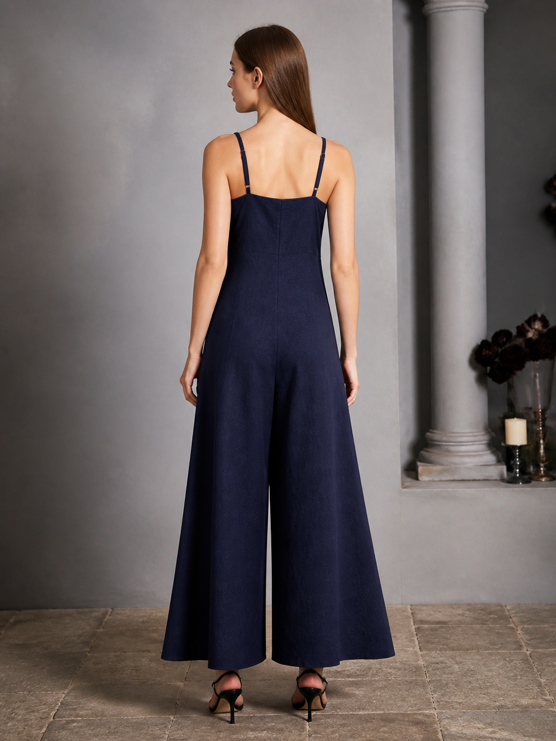Back view of a navy blue wide-leg jumpsuit with thin straps and flowing wide legs shown on a model indoors