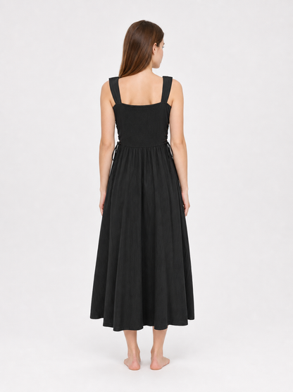 Back view of a black midi dress on a model, showing its square back neckline, broad straps, side ties, and gathered skirt