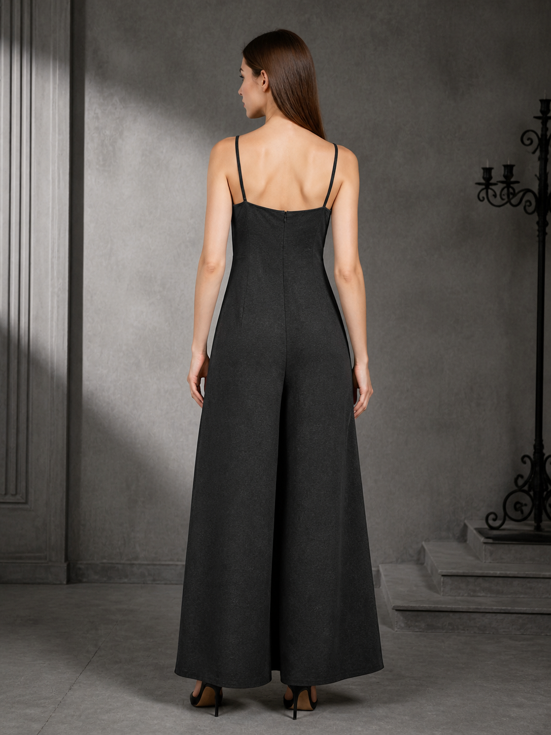 Back view of a black wide-leg jumpsuit showing thin straps, a fitted upper section, and flowing wide legs against a dark interior