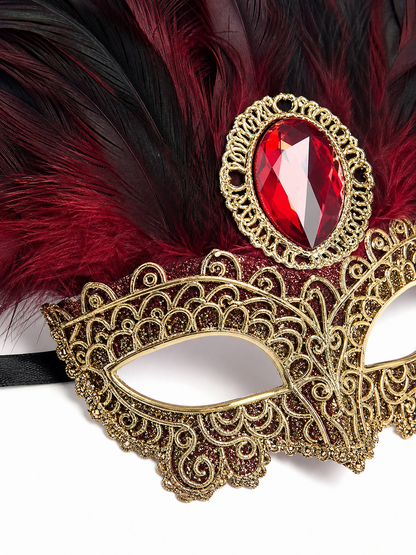Close-up of gold filigree mask detailing, red oval jewel, and layered burgundy and black feathers