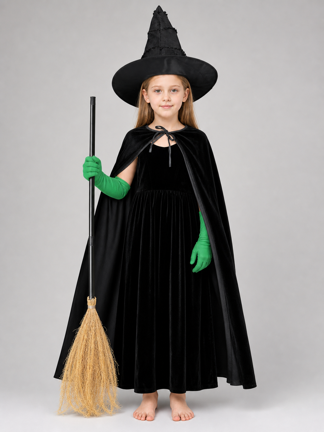 Full-body child model wearing a black velvet dress, cape, pointed hat, and green gloves, holding a broom and standing barefoot