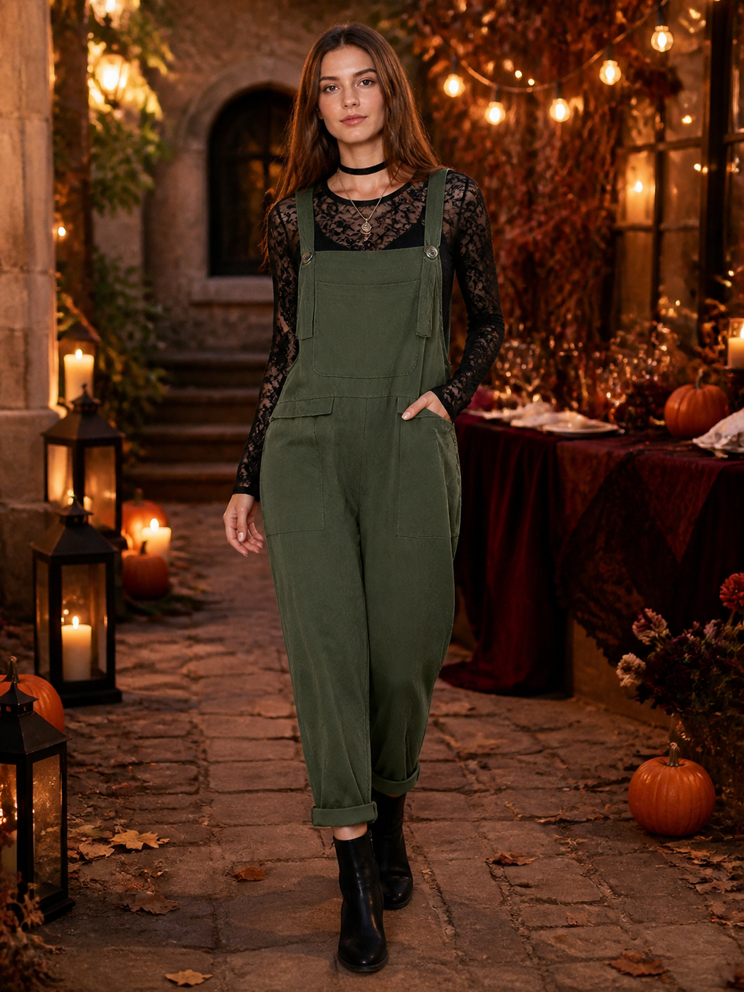 Full-body model wearing dark green bib overalls over a black lace top, with side pockets and rolled cuffs in a candlelit setting