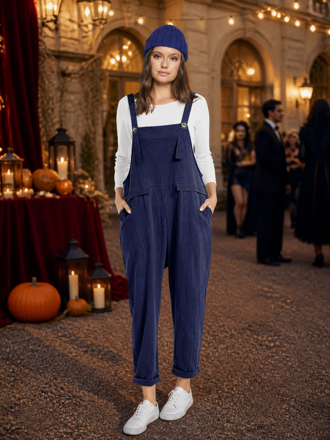 Navy blue bib overalls worn with a white shirt and blue knit hat in an indoor event setting, with white sneakers and cuffed hems