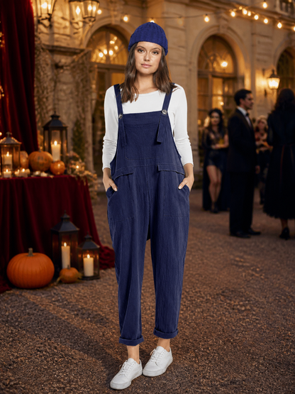 Navy blue bib overalls worn with a white shirt and blue knit hat in an indoor event setting, with white sneakers and cuffed hems