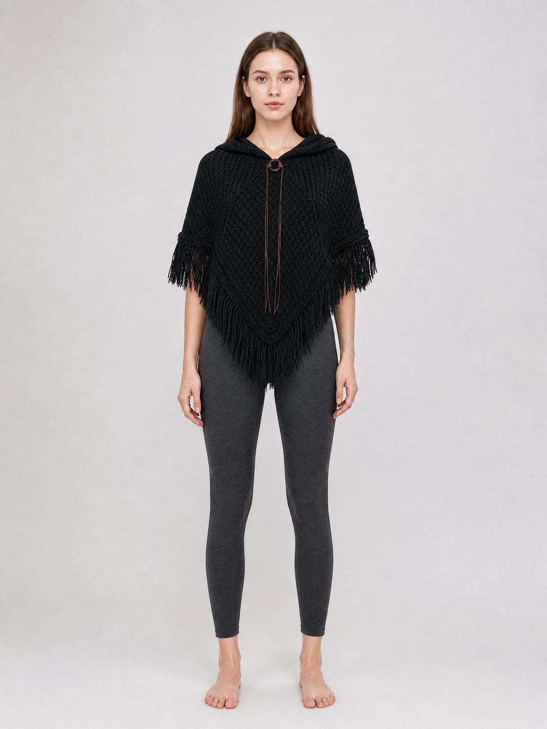 Black hooded knit poncho with long fringe, shown front-facing on a model wearing leggings against a gray background