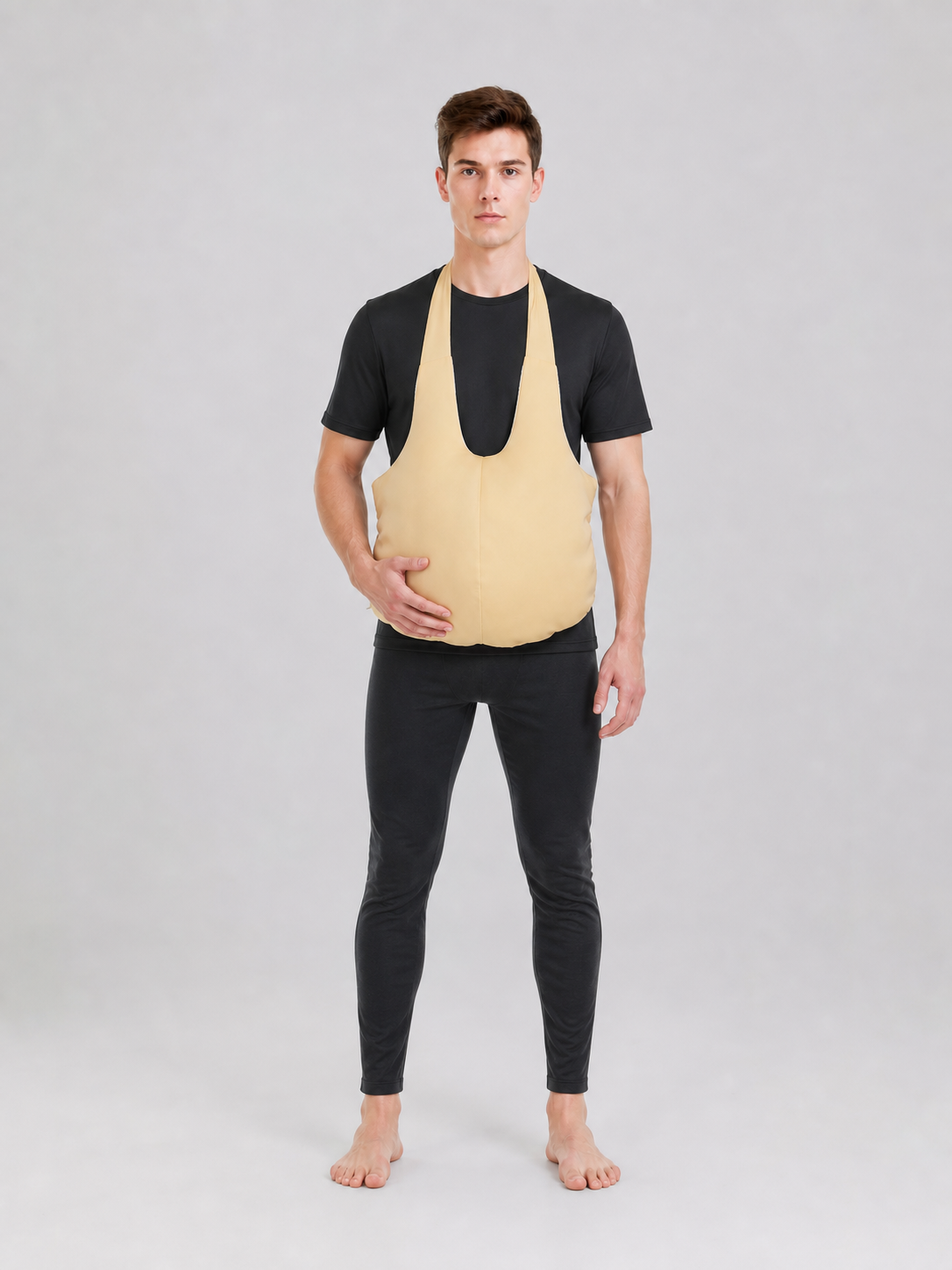 Wearable fake belly costume accessory with a large rounded shape. The model has a bib-style front panel, two shoulder straps, and side ties for fastening. The smooth solid-color surface comes in a light beige or beige shade, so the accessor