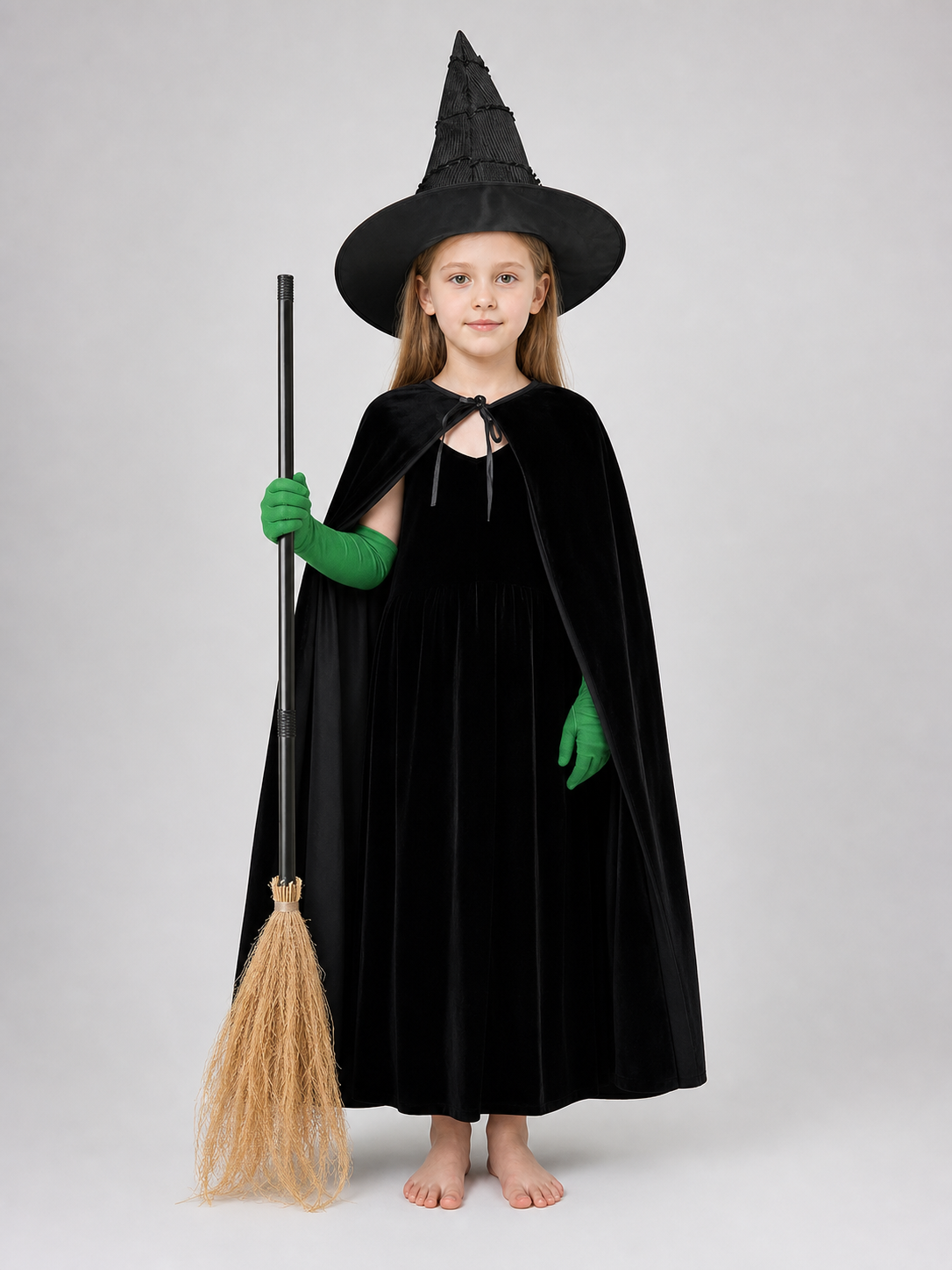 Full-body child model wearing a black dress, cape, and pointed hat with green gloves, holding a broom and standing barefoot