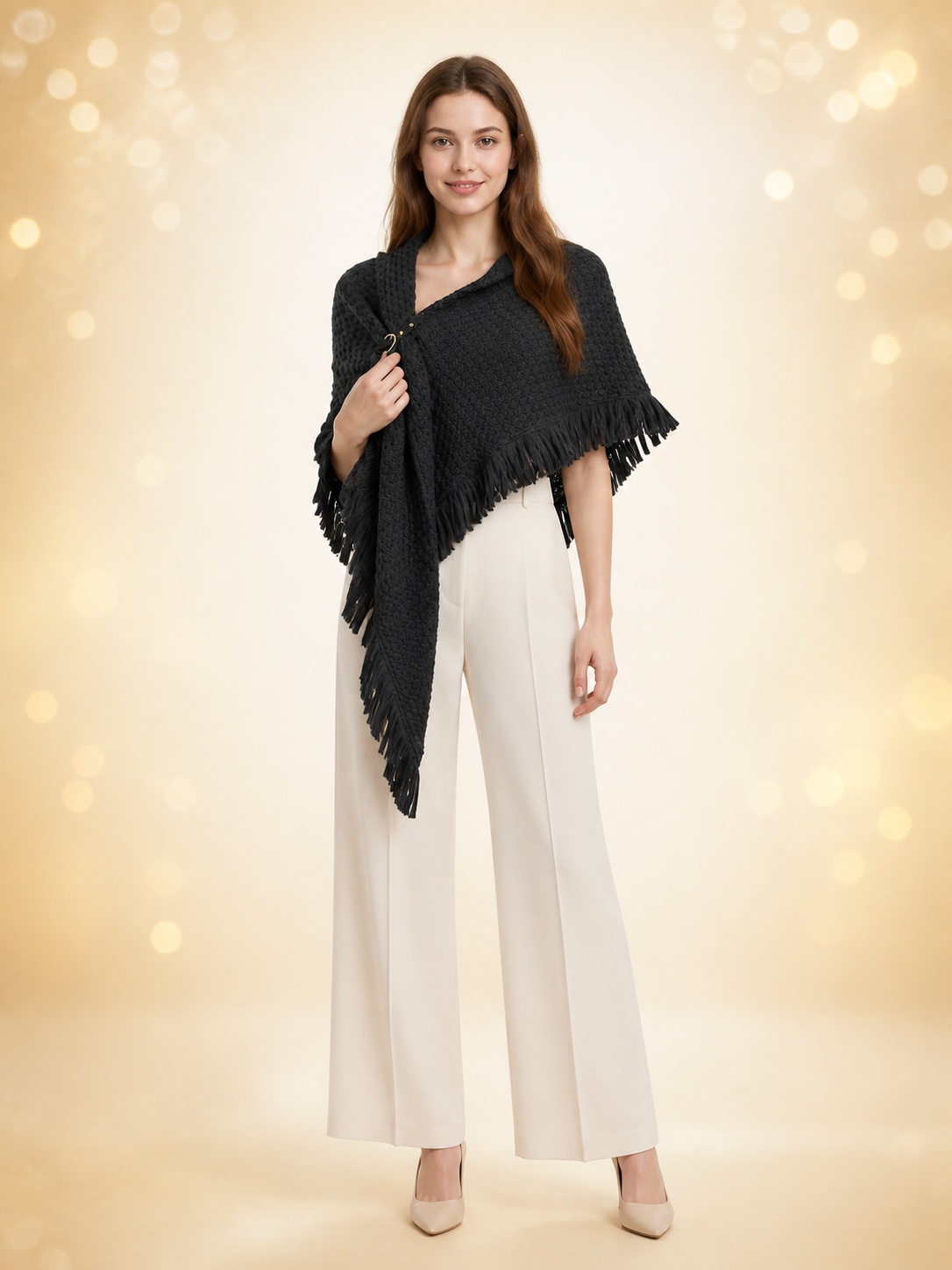 Black knitted poncho cape modeled full-body over white trousers, showing the triangular shape, fringe edging, and front ring closure