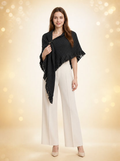 Black knitted poncho cape modeled full-body over white trousers, showing the triangular shape, fringe edging, and front ring closure