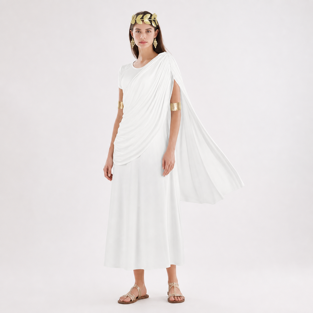 Three-quarter model view of a white draped dress with a shoulder train, gold arm bands and flat sandals against a plain background
