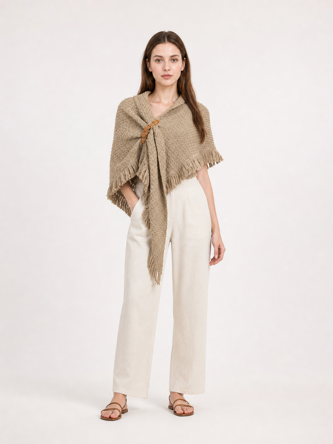 Full-body model wearing a beige woven cape with a long pointed front, side clasp, and fringe over white trousers