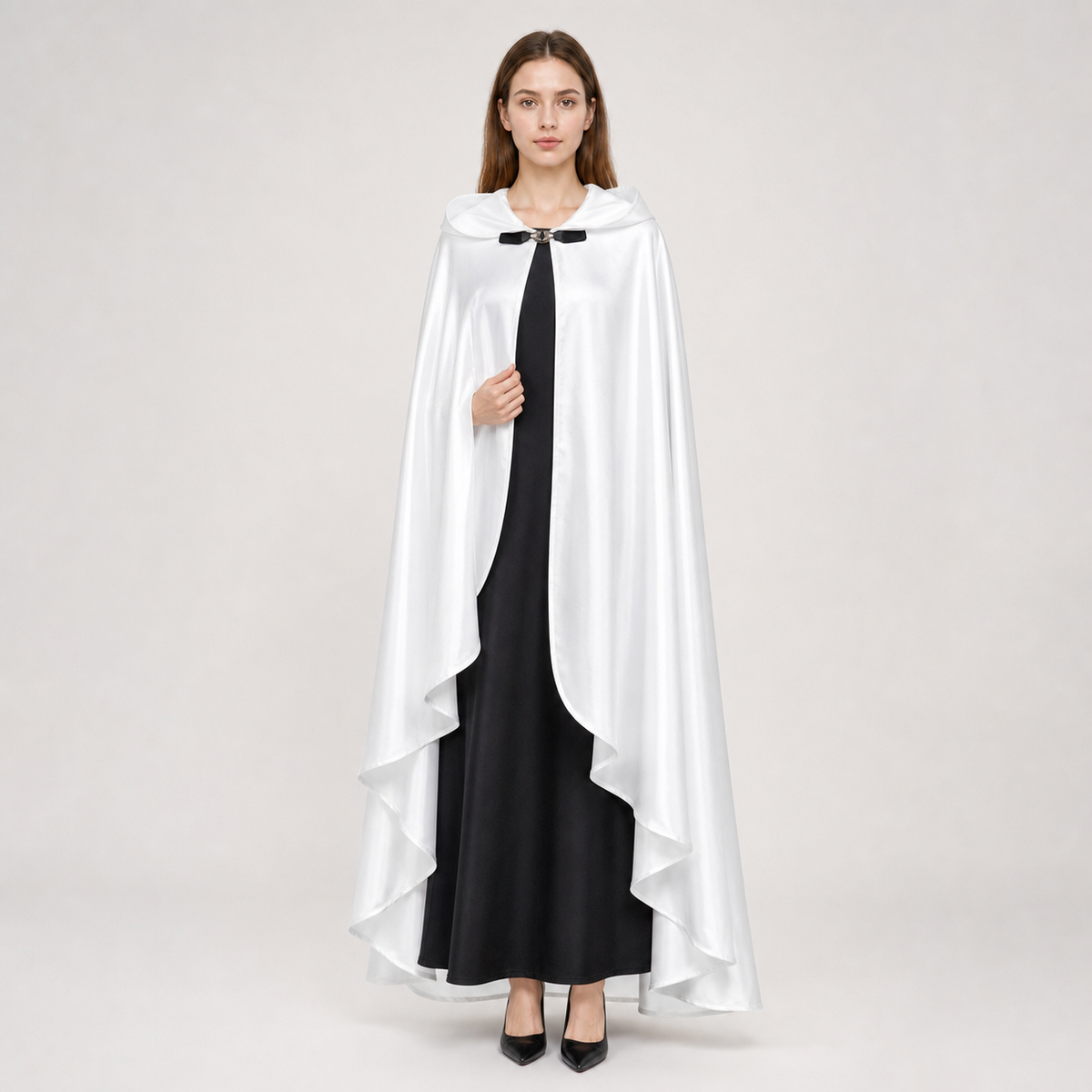 White hooded cape with glossy fabric, curved front opening, layered pointed hem, and black toggle closure, modeled over a long black dress