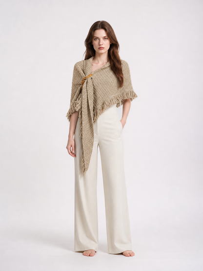 Beige woven cape with fringed edges and a side clasp, shown full-body over wide-leg white trousers on a studio model