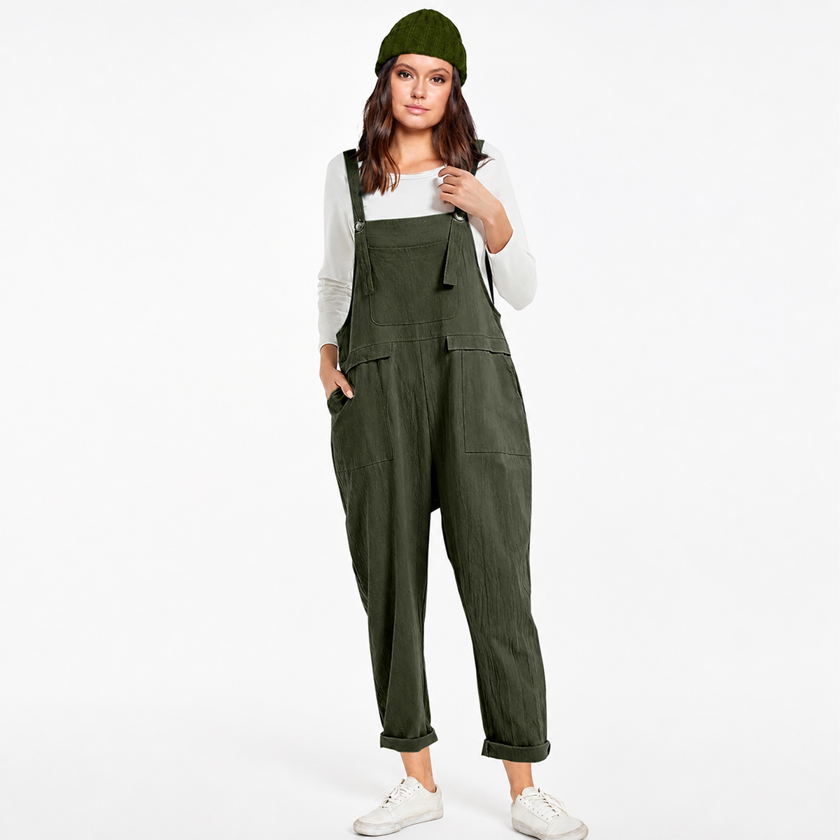 Full-body model wearing dark green bib overalls with a chest pocket, side pockets, rolled cuffs, white sneakers, and a matching knit hat