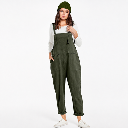 Full-body model wearing dark green bib overalls with a chest pocket, side pockets, rolled cuffs, white sneakers, and a matching knit hat