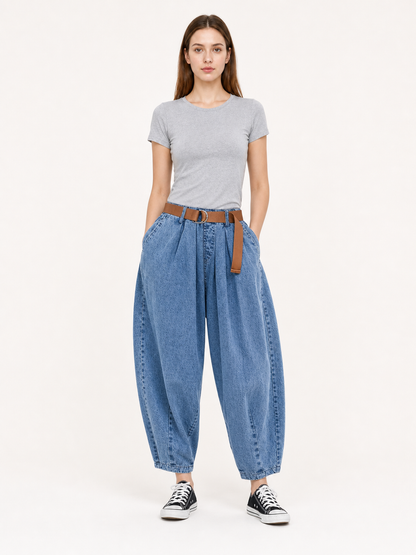 Front view of a model wearing blue denim pants with pleated wide legs, side pockets, a brown belt, and black sneakers